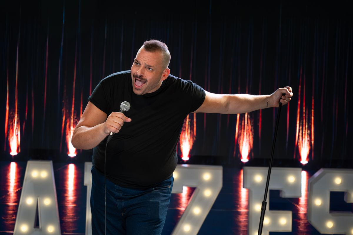 Daniel Franzese leans into the camera mid-joke, microphone in hand, in front of glowing marquee letters
