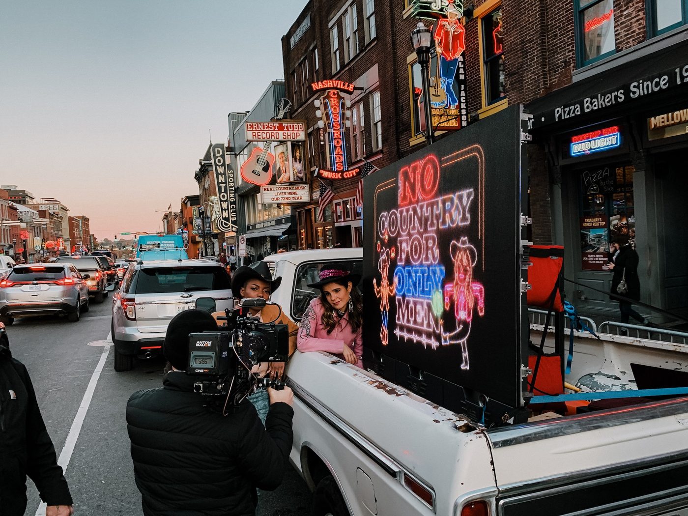 A Chronicle camera operator films two performers leaning from a pickup truck with a neon No Country For Only Men LED sign, on Broadway in Nashville