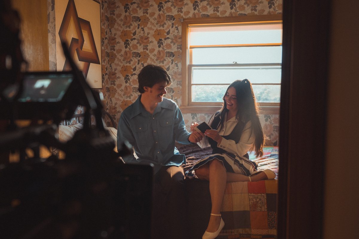 A young man and woman laugh together on a vintage bed, sharing a Polaroid in warm light.