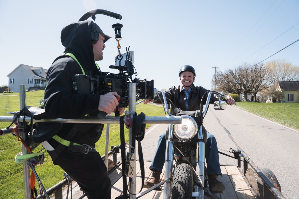 A camera operator films a motorcycle rider from a DIY process trailer on the road.